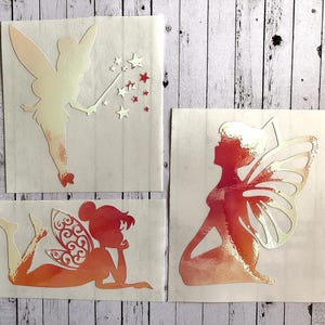 May include: Three fairy silhouette decals with a gradient of pink and orange. The fairies are in different poses, one is flying with a wand, one is sitting with wings spread, and one is lying down with wings folded.