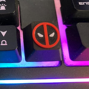 Puede incluir: Un teclado de computadora negro con teclas personalizadas, incluyendo una con un logotipo amarillo de Batman y otra con un icono de máscara de Deadpool en rojo y negro, sobre un fondo de efectos de iluminación RGB multicolor integrados.