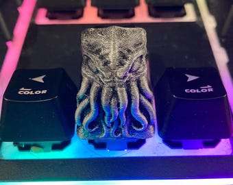 May include: A custom gray Cthulhu keycap sits between two black keyboard keys with "COLOR" text. The keyboard has a black frame and is illuminated with a rainbow of colors.