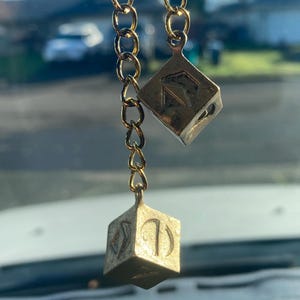 May include: Two gold-colored, cube-shaped charms hang from a gold chain. The charms have etched symbols and numbers on their faces. The background is blurred, showing a sunny outdoor scene with houses and trees.