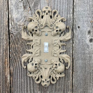 May include: A beige, decorative light switch cover with a skull and tentacle design. The cover has a rectangular shape with a single light switch in the center. The design includes multiple skulls and ornate detailing. The cover is mounted on a weathered wooden surface.