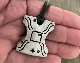 May include: A silver-colored pendant with a black cord. The pendant has a unique shape with curved lines and small circular and diamond-shaped details. The pendant is held in a hand.