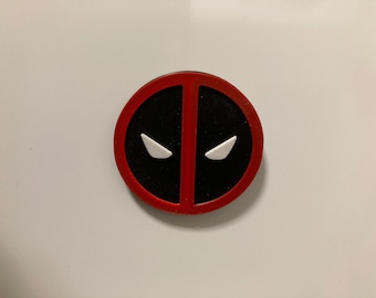 May include: A circular magnet featuring the Deadpool logo. The design consists of a red outer ring and a vertical red bar dividing a black background. Two white, angular, eye-shaped cutouts are positioned symmetrically within the black sections.