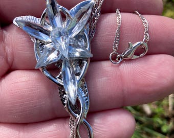 May include: A silver-toned necklace featuring a star-shaped pendant with clear crystal accents. The pendant is suspended from a delicate chain with a lobster clasp. The necklace is displayed against a blurred green background.