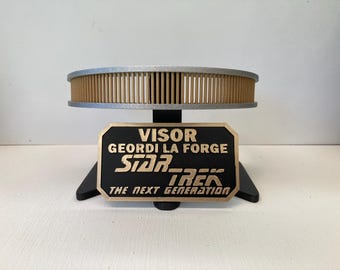 May include: A black and gold Star Trek Geordi La Forge visor display stand. The stand features the text "VISOR GEORDI LA FORGE STAR TREK THE NEXT GENERATION". The visor is a light brown color with vertical slats.