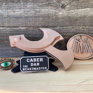 May include: A collection of copper-colored fantasy-themed items, including a stylized axe, a circular emblem with a bird design, and a decorative eye pin. A black sign reads "Caber the Beastmaster."