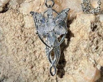 May include: A silver-colored pendant necklace featuring a stylized floral design with clear, faceted crystals. The pendant has a detailed, ornate frame and hangs from a delicate chain. The necklace is displayed on a textured, light-colored stone surface.