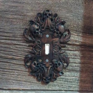 May include: A decorative light switch cover with a gothic design featuring skulls and vines. The cover is made of a dark brown material and has a white light switch.