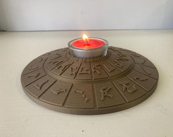 May include: A brown, circular candle holder with an intricate, engraved design. A lit red candle sits in a clear glass holder in the center. The candle holder is a decorative item.