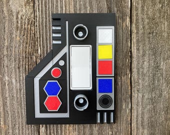 May include: Black rectangular panel with various colored buttons and dials. Features a white rectangle, a series of colored squares (white, yellow, red, blue), and two circular dials. The panel has a geometric design with silver and gray accents.
