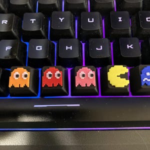 Könnte beinhalten: Nahaufnahme einer schwarzen Tastatur mit individuellen Tastenkappen, die pixelige Pac-Man-Charaktere in Orange, Rot, Rosa, Gelb und Blau zeigen. Die Tastenkappen sind in einer Reihe angeordnet.