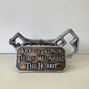 May include: A silver-colored key with a rectangular plaque featuring the text "KEY TO EREBOR THORIN OAKENSHIELD THE HOBBIT." The key has a decorative design with geometric shapes and is likely a prop or collectible item.