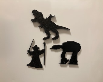 May include: Three black glittery silhouettes of fictional characters and a vehicle. One is a dinosaur with a rider, one is a robed figure holding a staff and sword, and the third is a four-legged mechanical walker with a flat top and angular body.