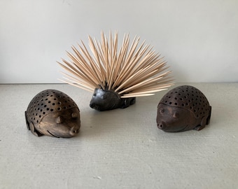 May include: Three decorative hedgehog figurines. Two are brown with small holes, and one is black with wooden toothpicks as quills. The figurines are arranged on a light-colored surface.