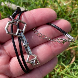 May include: A silver-colored pendant in the shape of a key, suspended on a black cord necklace. The key has geometric cutouts and decorative details. The necklace is held in a hand against a backdrop of green grass.
