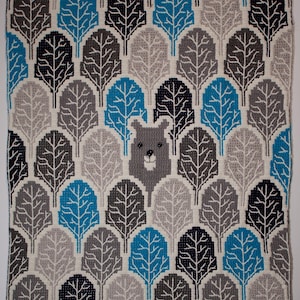Könnte beinhalten: Eine gehäkelte Decke mit einem Muster aus Bäumen in Grautönen, Blau und Schwarz. Ein Bärenkopf befindet sich in der Mitte der Decke.
