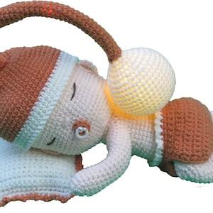 May include: A crocheted baby doll with a brown hat and a white nightgown is sleeping on a white pillow. The doll has a light-up bulb attached to its head.
