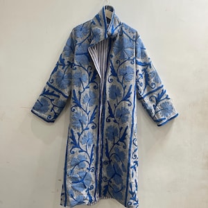 May include: A long-sleeved coat featuring a light gray base with vibrant blue floral embroidery throughout. The garment has a high collar, an open front design, and a visible striped lining on the inner edges. The embroidery consists of large flower motifs and swirling vine patterns covering the entire exterior surface.