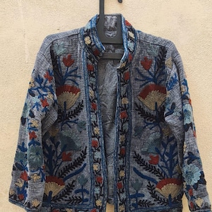 May include: A blue textured jacket featuring a high collar and long sleeves, adorned with intricate floral embroidery in shades of red, yellow, and light blue. The garment has a front-opening design with a dark blue trim along the edges.