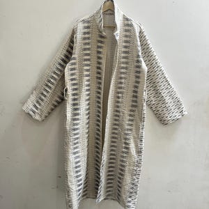 May include: A long-sleeved, open-front coat featuring a cream-colored base with vertical stripes in shades of charcoal gray. The garment is constructed with visible running stitch detailing throughout the fabric, creating a textured, quilted appearance.