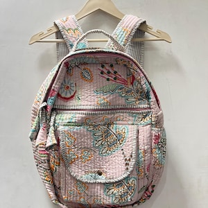 May include: A quilted backpack in a soft pink hue featuring a multi-colored floral and botanical print. The bag includes a top handle, two adjustable shoulder straps, a main zippered compartment, and a front pocket with a single snap closure.