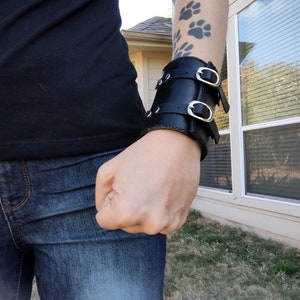 May include: Black leather wrist cuff with two silver buckles. The cuff is worn on a person's wrist, and the design includes two straps with silver buckles. The person is wearing a black shirt and blue jeans.