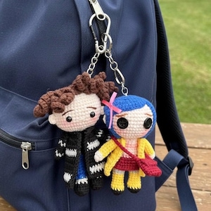 May include: Two crocheted doll keychains hang from a navy blue backpack. One doll has brown hair and a black coat, the other has blue hair, a yellow coat, and a red purse. The dolls are attached to the backpack with silver chains.