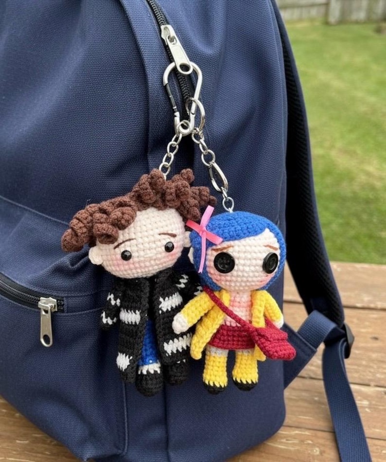 May include: Two crocheted doll keychains hang from a navy blue backpack. One doll has brown hair and a black coat, the other has blue hair, a yellow coat, and a red purse. The dolls are attached to the backpack with silver chains.