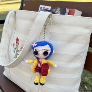 May include: A handmade crochet doll keychain with blue hair, a yellow jacket, and a red bag, hanging from a cream-colored tote bag. The tote bag has a floral embroidery design and is filled with books. The keychain is attached to the bag's strap.