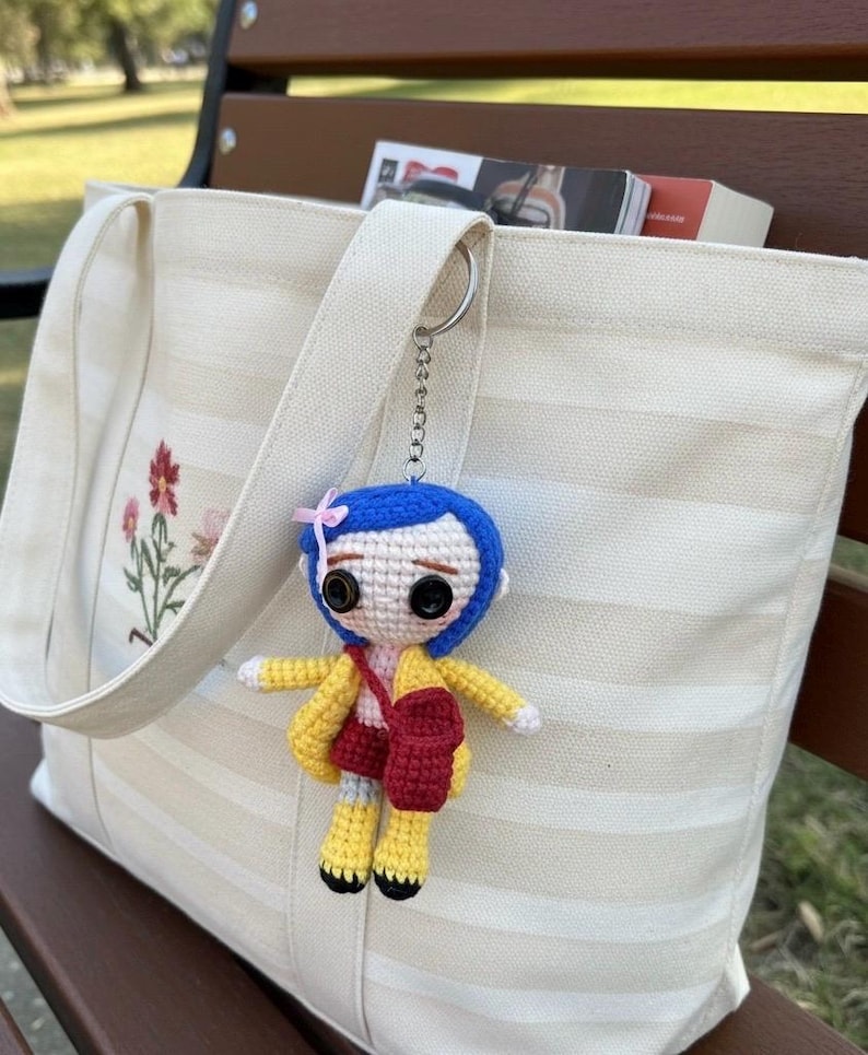 May include: A handmade crochet doll keychain with blue hair, a yellow jacket, and a red bag, hanging from a cream-colored tote bag. The tote bag has a floral embroidery design and is filled with books. The keychain is attached to the bag's strap.