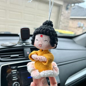 May include: A handmade crochet doll in a seated pose, featuring a yellow top, black hair, and a pink and white lotus flower base. The doll has a red dot on its forehead and is suspended by a white string, suitable for hanging as a car charm or decorative item.