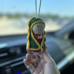 May include: A crocheted figurine of the Virgin Mary, wearing a green and yellow robe, hanging from a rearview mirror in a car.