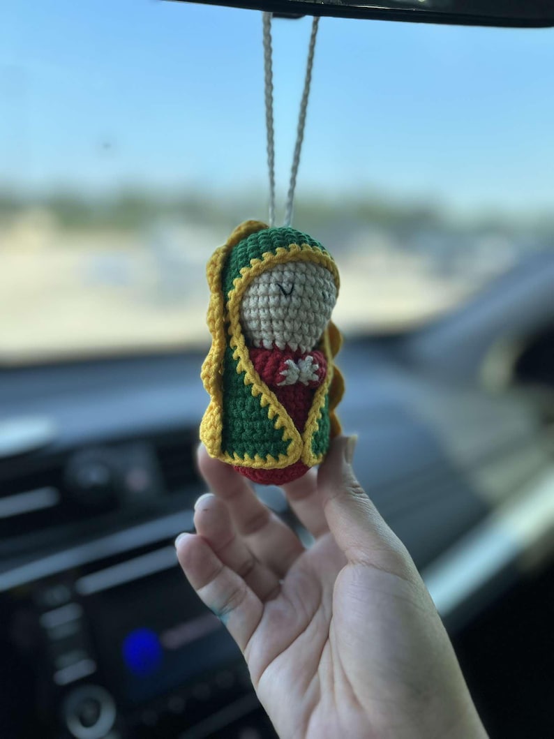 May include: A crocheted figurine of the Virgin Mary, wearing a green and yellow robe, hanging from a rearview mirror in a car.
