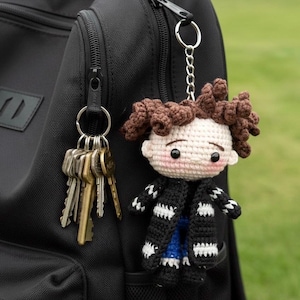 May include: A handmade crochet doll keychain with brown hair, a black and white striped coat, and a blue shirt. The doll is attached to a keyring with keys, hanging from a black backpack. The background is blurred green grass.