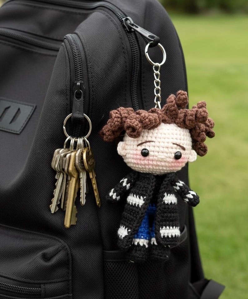 May include: A handmade crochet doll keychain with brown hair, a black and white striped coat, and a blue shirt. The doll is attached to a keyring with keys, hanging from a black backpack. The background is blurred green grass.