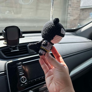May include: A crocheted black and white Batman character hanging from a rearview mirror in a car. The character has a yellow belt and a black cape.
