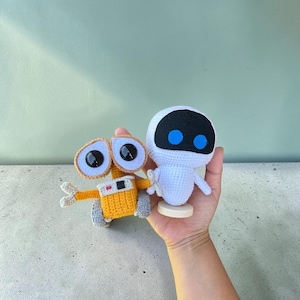 May include: Two handmade crochet dolls representing robotic characters. One is a yellow and grey rectangular robot with large black eyes, and the other is a white, rounded robot with a black face panel and two bright blue circular eyes.