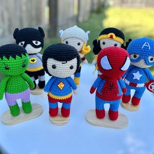 May include: A collection of seven handmade crochet superhero dolls. Each doll is approximately 6 inches tall and features vibrant colors and iconic details. Characters include Batman, Superman, Captain America, Spider-Man, Hulk, Thor, and Wonder Woman.