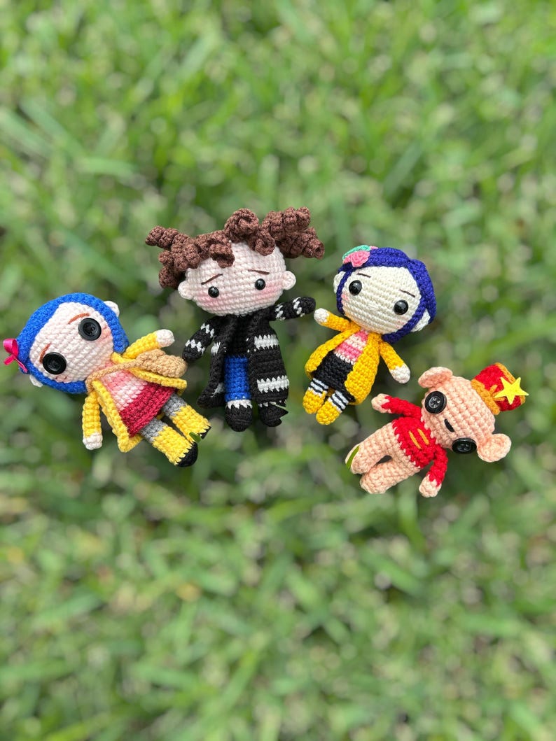 May include: Four handmade crochet dolls, each with unique outfits and features. One doll has blue hair and a yellow coat, another has brown hair and a black coat, and two others have red and yellow outfits. They are arranged on a green grassy surface.