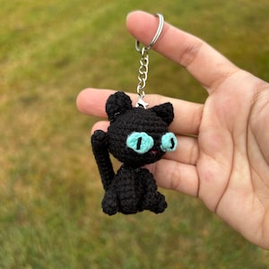 May include: A black crocheted cat keychain with bright blue eyes and a silver keyring. The handmade cat has a small tail and is held up against a blurred green background. The cat's eyes are outlined in black and have a red mouth.