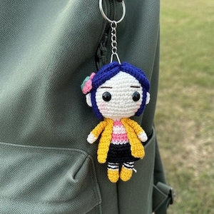 May include: Crocheted doll keychain featuring a character with blue hair, a yellow jacket, and black shorts. The doll has a pink and white striped shirt and is attached to a silver keyring. The keychain is hanging on a green backpack.