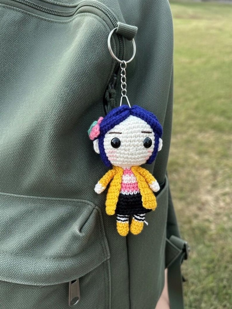May include: Crocheted doll keychain featuring a character with blue hair, a yellow jacket, and black shorts. The doll has a pink and white striped shirt and is attached to a silver keyring. The keychain is hanging on a green backpack.