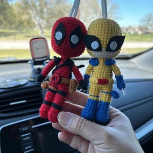 May include: Two crocheted superhero figures hanging from a rearview mirror. The figure on the left is red and black and resembles Deadpool. The figure on the right is yellow and blue and resembles Wolverine.