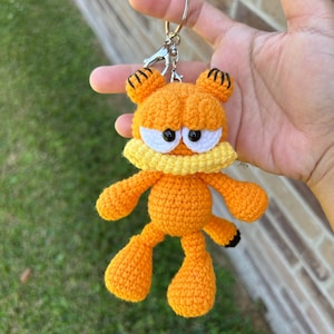 May include: Orange crochet Garfield keychain with black eyes, a yellow collar, and black stripes on the ears. The handmade keychain is attached to a silver keyring.