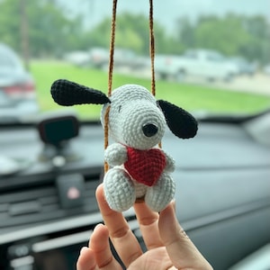 May include: A small, handmade crochet ornament featuring a light gray dog with long black ears and a black nose, holding a bright red heart. The figure is suspended by a thin brown cord, designed to hang as a decorative accessory.