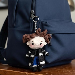 May include: A handmade crochet doll keychain with brown curly hair, a black and white striped scarf, and blue jeans. The doll is attached to a silver keyring and hangs in front of a navy blue backpack. The doll has a light skin tone and rosy cheeks.
