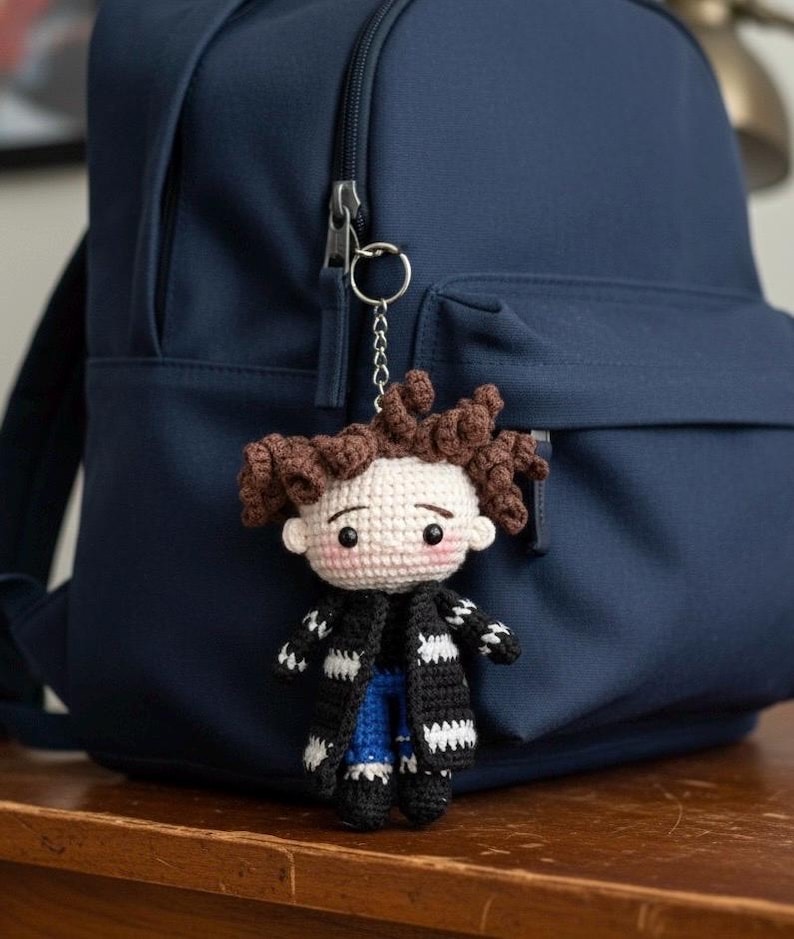May include: A handmade crochet doll keychain with brown curly hair, a black and white striped scarf, and blue jeans. The doll is attached to a silver keyring and hangs in front of a navy blue backpack. The doll has a light skin tone and rosy cheeks.