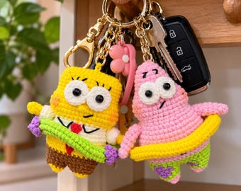 May include: Two crocheted keychains featuring yellow square and pink starfish characters. The yellow character wears a white shirt, red tie, and brown pants, holding a purple object. The pink character wears green shorts with purple flowers.