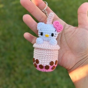 May include: Crocheted keychain featuring a white cat character with a pink bow, sitting in a pink and brown boba drink cup. The keychain is attached to a silver metal ring.