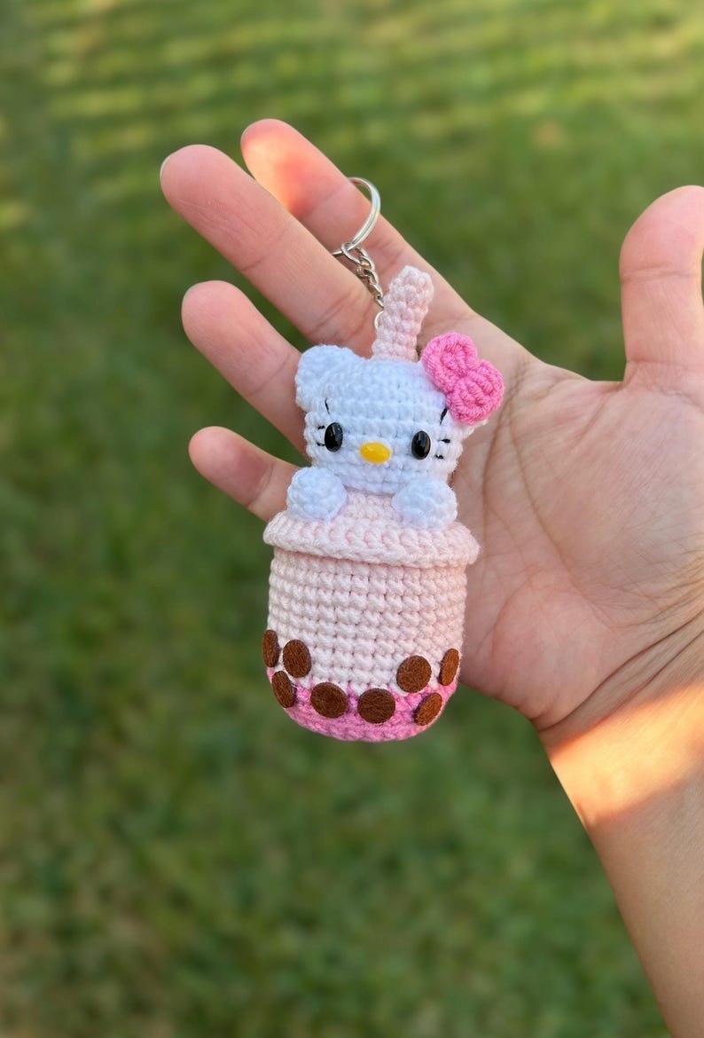 May include: Crocheted keychain featuring a white cat character with a pink bow, sitting in a pink and brown boba drink cup. The keychain is attached to a silver metal ring.